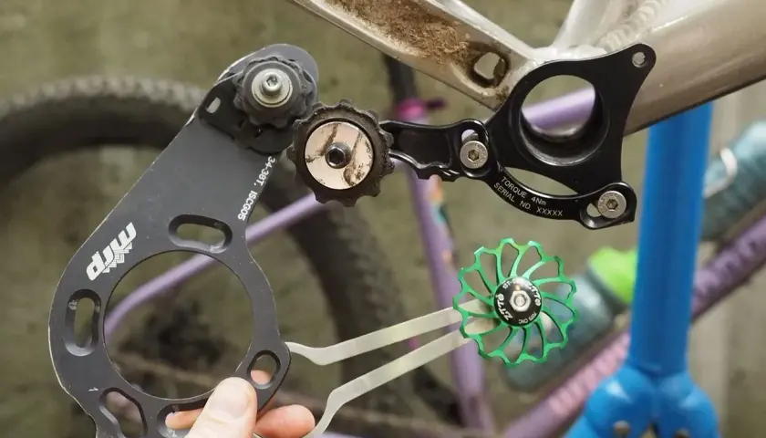 A hand holds a bike chain guide, part of a 1x drivetrain, near a bicycle frame. A green pulley wheel is also visible.