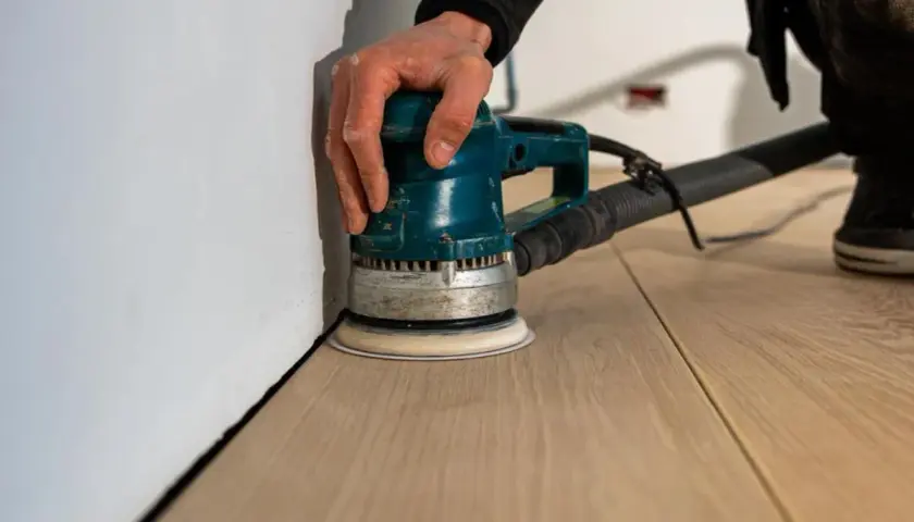 Mano che usa una levigatrice orbitale per levigare il parquet fai da te, avvicinandosi al battiscopa bianco.