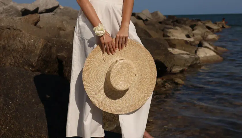 Mujer con un sombrero para ir a los toros, vestida de blanco, de pie sobre rocas junto al mar.