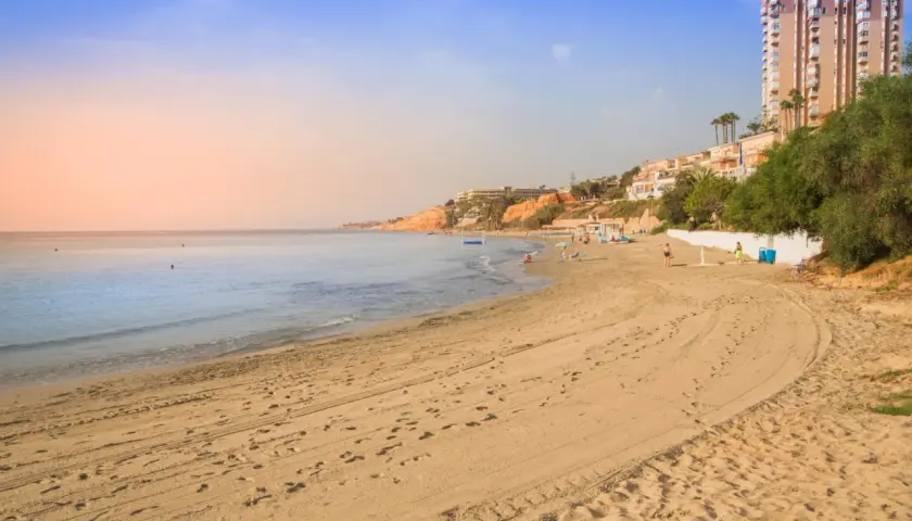 Playa con arena dorada y el mar azul. Al fondo, un barranco rubio con edificios y vegetación.