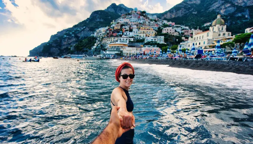Kobieta w czarnym stroju kąpielowym trzyma za rękę, prowadząc na **neapolską plażę** z widokiem na kolorowe domy Positano.