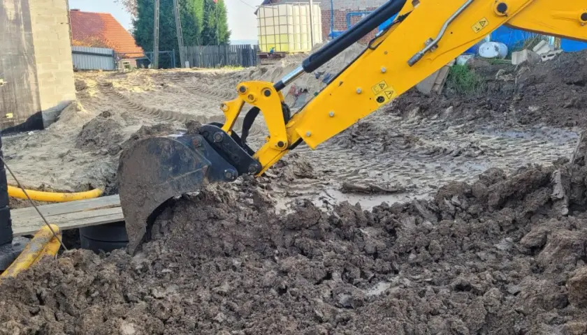 Żółta koparka marki JCB pracuje na budowie, kopiąc ziemię. Widać jej ramię i łyżkę zagłębiającą się w błotniste podłoże.