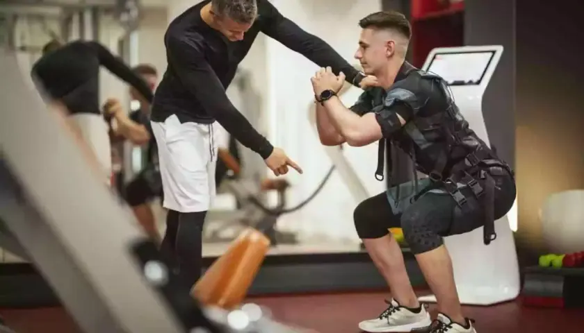 Trener instruuje mężczyznę w kamizelce EMS podczas ćwiczeń w siłowni.