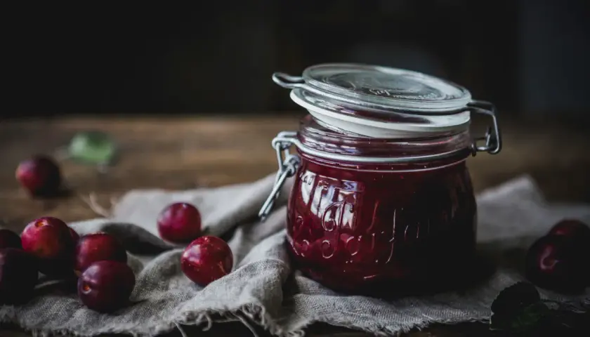 En burk med hemlagad plommonmarmelad med kanel, omgiven av färska plommon på en linneduk.