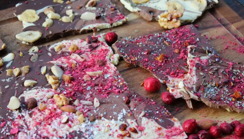 Bruchschokolade selber machen: Bunte Schokoladenstücke mit Nüssen, Beeren und Bananenchips auf Holzbrett.