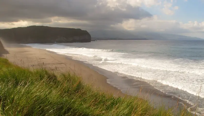 Ein einsamer azoren strand mit sanften Wellen und dramatischen Wolken. Hohes Gras wiegt sich im Wind.