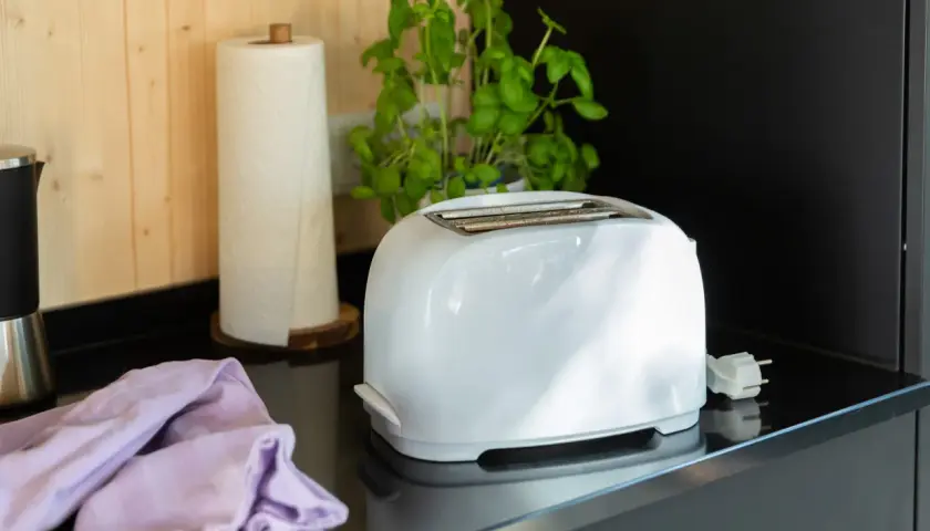 Ein weißer Toaster steht auf einer schwarzen Arbeitsplatte, bereit für die Reinigung. Daneben liegen ein lila Tuch und eine Küchenrolle.