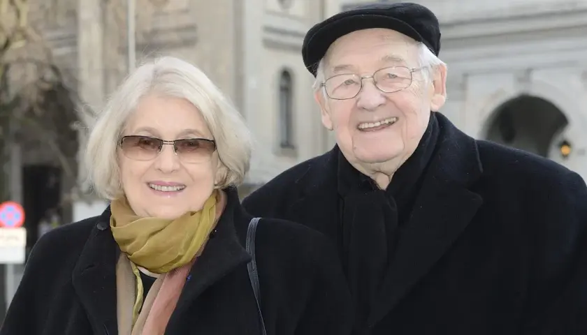 Uśmiechnięta para: Andrzej Wajda i jego żona, w eleganckich płaszczach i szalikach, pozują do zdjęcia na tle architektury.