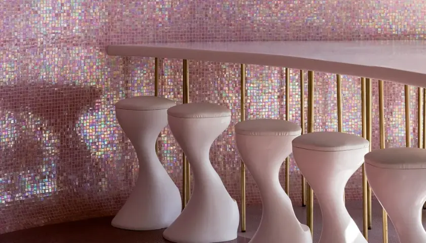 Un bancone rosa pallido con sgabelli bianchi si staglia contro un muro scintillante di **bisazza ceramiche** iridescenti, creando un'atmosfera glamour.