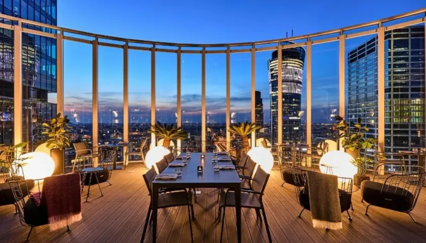 Przytulna restauracja Warszawa na dachu z widokiem na nocne miasto. Stoły i krzesła, palmy i kule świetlne tworzą magiczną atmosferę.