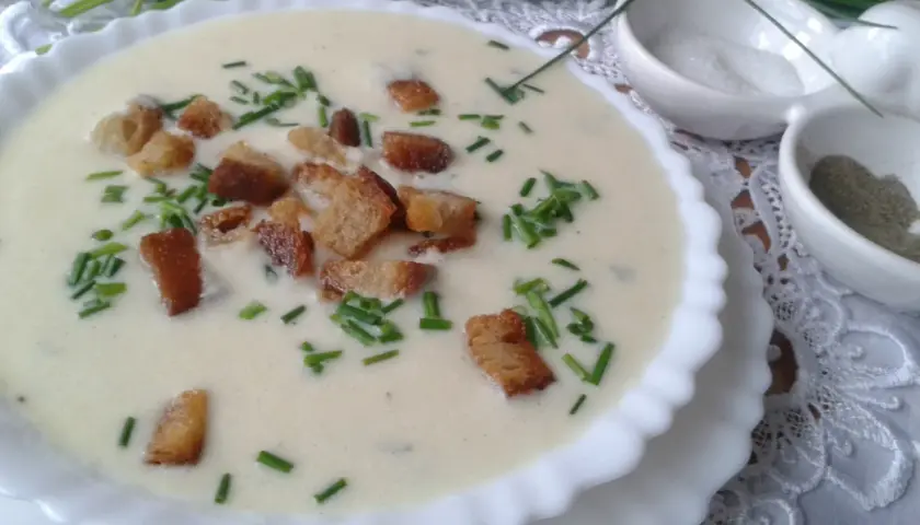Kremowa zupa porowa z grzankami i szczypiorkiem, podana w białej miseczce. Obok małe miseczki z solą i pieprzem.