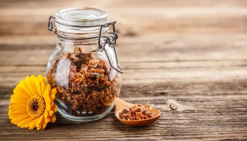 Propoli grezza in un barattolo di vetro, con un cucchiaino e un fiore giallo. Scopri come usarla per il benessere.