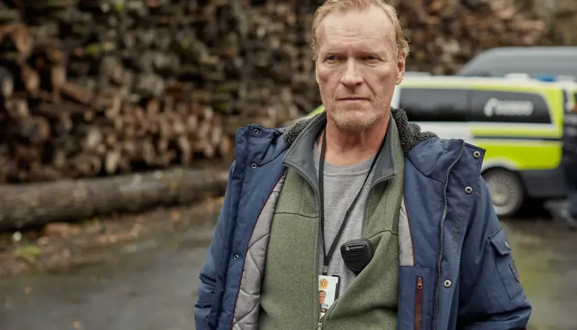 En man med intensiv blick, klädd i jacka och väst, bär en radio och ett id-kort. Bakom honom syns en polisbil och en vedhög. En scen från en norsk serie.