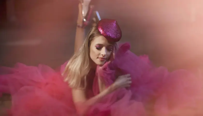 Urocza sesja glamour: blondynka w różowej tiulowej sukni i błyszczącej beretce, z makijażem oczu w tym samym kolorze, leży na ziemi.