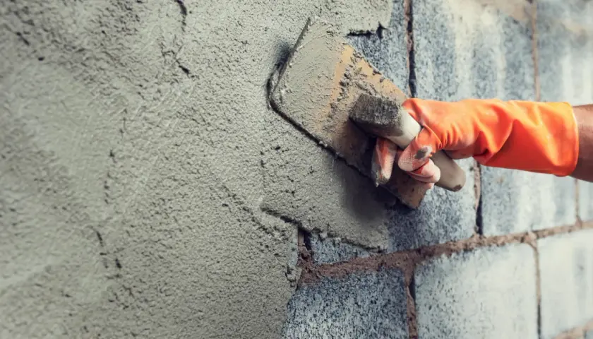 Mano con guanto arancione stende malta per intonaco esterno su un muro di blocchi di cemento con una cazzuola.