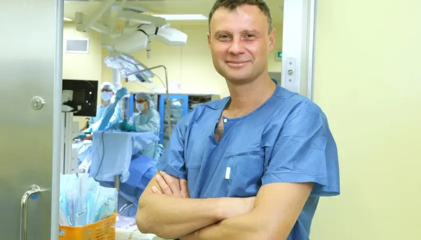 Lekarz w niebieskiej koszuli chirurgicznej stoi na tle sali operacyjnej.