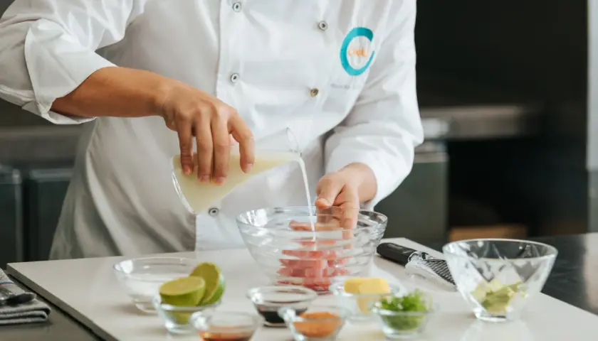Chef vierte líquido en un bol con pescado, preparando la leche de tigre receta. Ingredientes frescos como limas y aguacate a la vista.