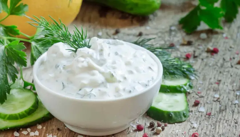 Pyszny dip jogurtowy z ziołami i ogórkiem, udekorowany koperkiem, podany z plastrami ogórka i przyprawami na drewnianym blacie.