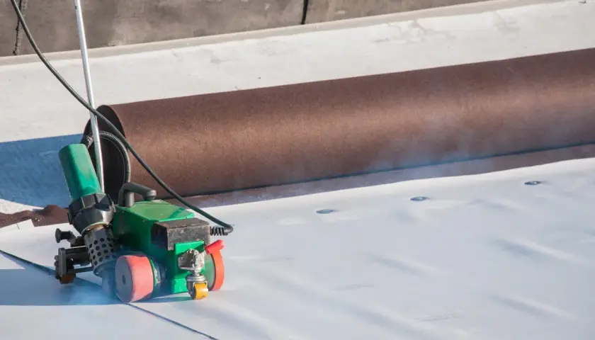 Gorące powietrze zgrzewarki topi białą membranę EPDM, zapewniając jej szczelny montaż. Obok zwinięta brązowa rolka materiału.