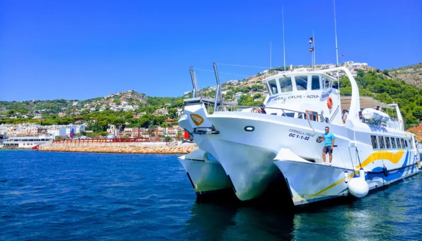 Catamarán "Estrella Fugaz" en el puerto de Dénia, con la costa y el monte al fondo.