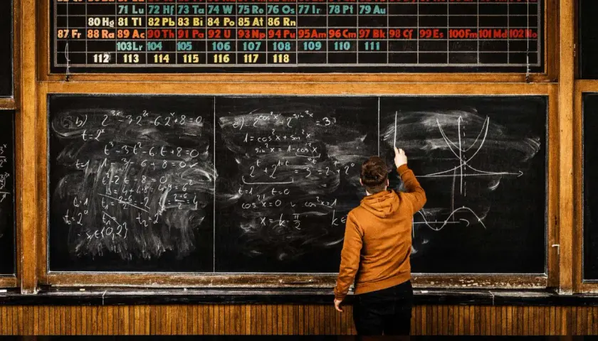 Chłopak na rok przerwy (gap year) rozwiązuje zadania z matematyki i fizyki przy tablicy, nad którą wisi układ okresowy pierwiastków.