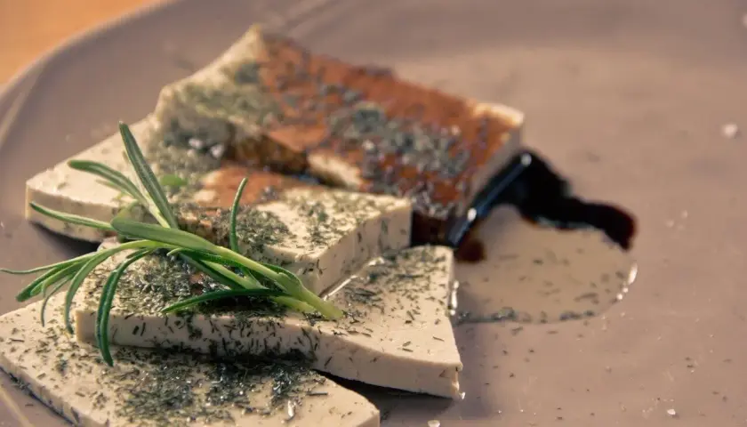 Queso tofu marinado con hierbas y romero, servido en un plato con salsa oscura.
