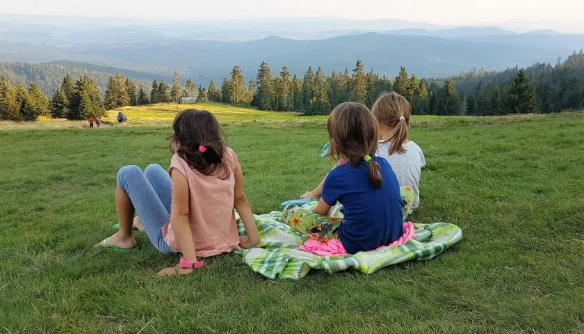 Trzy dziewczynki, jak małe rysianki, siedzą na trawie, podziwiając widok gór. Jedna z nich, z żabnicą na głowie, wpatruje się w dal.