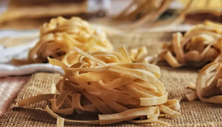 Nidi di tagliatelle fatte in casa, pronte per essere cucinate. La pasta fresca, con la sua texture rustica, è disposta su una tela grezza, invitando a un pasto autentico.