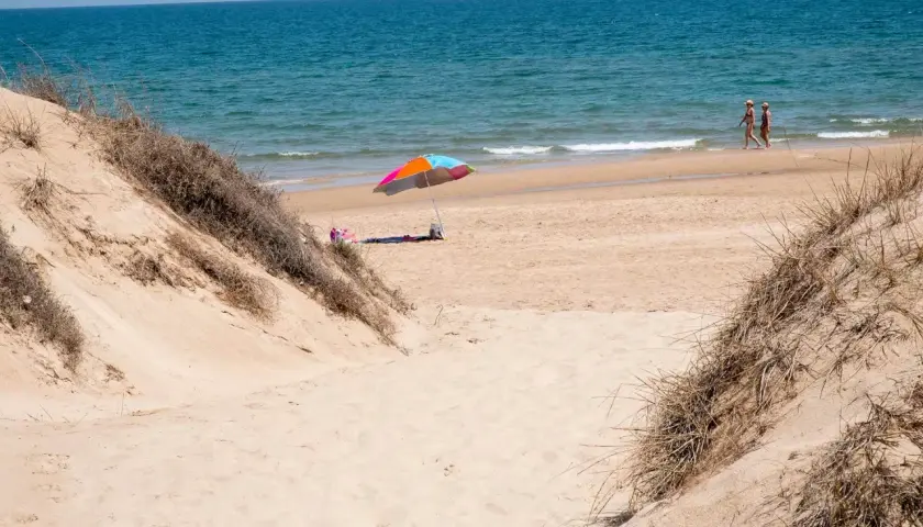 Dunas de arena y mar azul en el mareny de Sant Llorenç. Una sombrilla colorida y dos personas pasean por la orilla.
