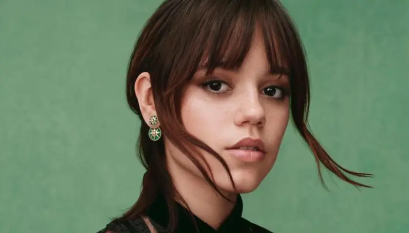 Jenna Ortega z kolczykami w kształcie gwiazdek. Jej włosy są ciemnobrązowe, a tło zielone.