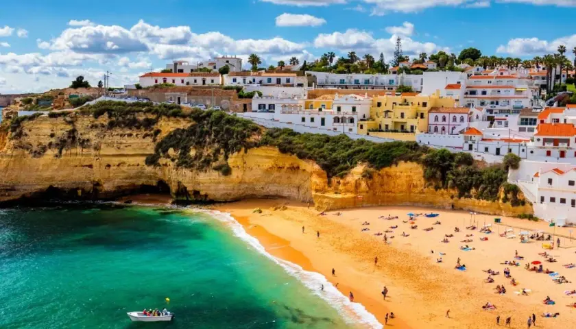 Sonnige Küste Portugals im Oktober. Ein Boot auf dem türkisfarbenen Meer vor einer Bucht mit Sandstrand und weiß-gelben Häusern.