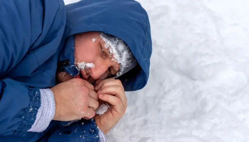 Mężczyzna w niebieskiej kurtce z kapturem, z twarzą pokrytą śniegiem, przytula dłonie. Czy to hipotermia, co to za stan?