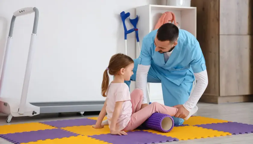 Fizjoterapeuta stosuje metodę Bobath podczas ćwiczeń z dziewczynką, która leży na macie z wałkiem pod nogą.