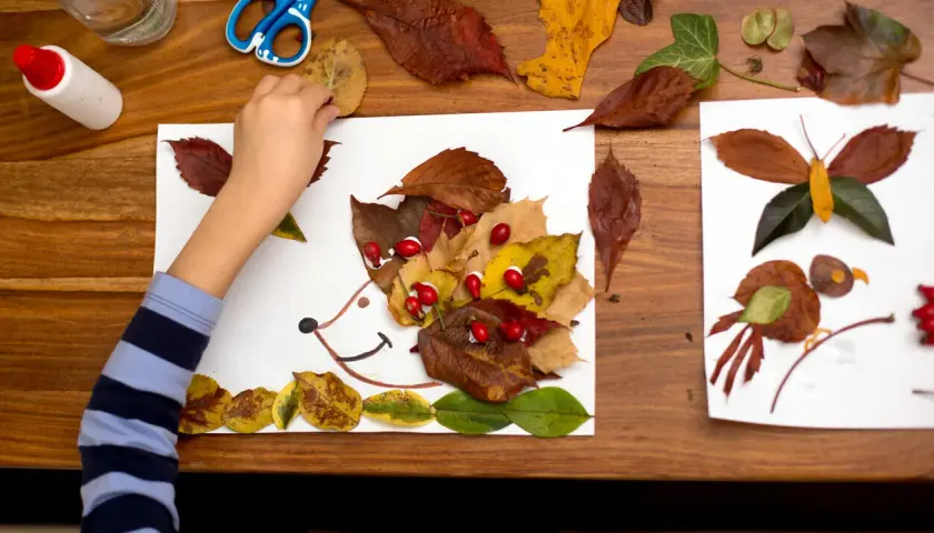 Niño haciendo otoño manualidades preescolar: un erizo con hojas secas y bayas rojas, junto a una mariposa y un pájaro hechos de hojas.