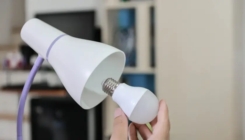 Ręka wymienia żarówkę w lampce biurkowej, symbolizując modernizację oświetlenia i oszczędność energii.