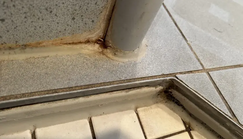 Schimmel in Fliesenfugen, oft durch Feuchtigkeit verursacht, zeigt sich als dunkle Verfärbungen. Hier sichtbar an einer Ecke mit Metallleiste.