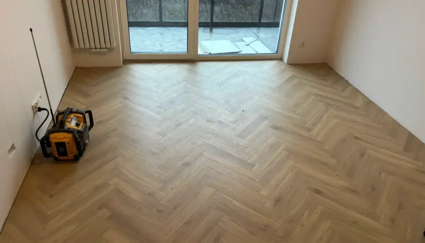 Nowy, drewniany panel winylowy ułożony we wzór jodełki. W rogu stoi żółto-czarne urządzenie.