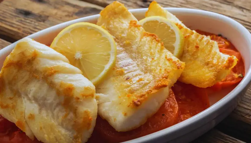 Filetes de pescado blanco horneado con rodajas de limón y tomates en una fuente blanca.