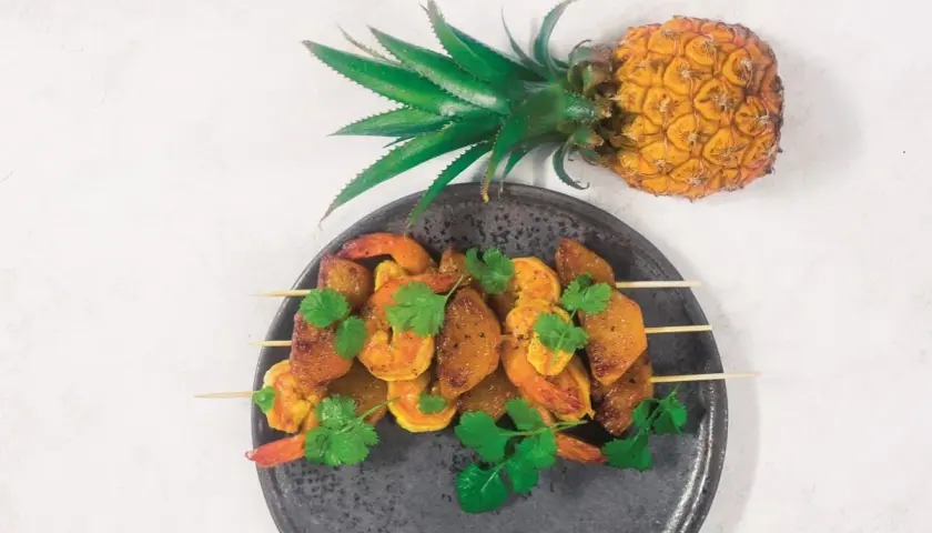 Szpadki z krewetkami i kawałkami ananasa, ozdobione kolendrą. Obok leży cały ananas, symbolizujący owoce egzotyczne.