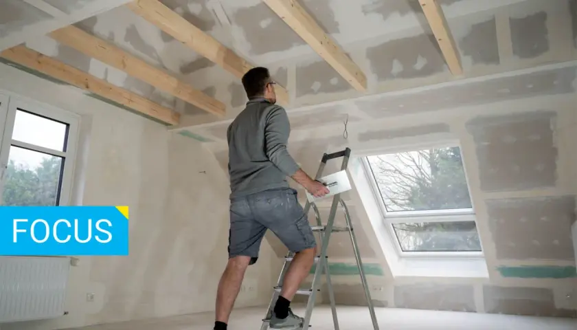Un operaio su una scala lavora al soffitto, applicando stucco su pannelli di fibrogesso, materiale versatile per ristrutturazioni.