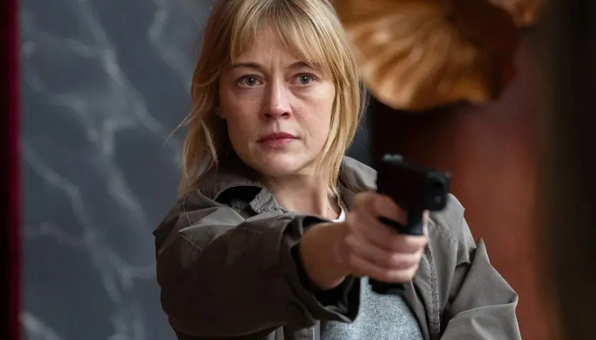 En kvinna med blont hår och en grå jacka håller en pistol redo. Hon är en del av rollistan i morden och ser beslutsam ut.