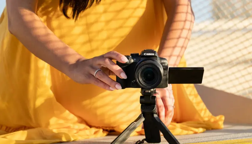 Kobieta w żółtej sukience ustawia aparat Canon EOS R10 na statywie, eksperymentując z trybem manualnym w aparacie.