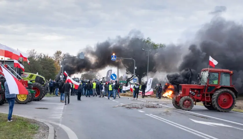 Czerwony traktor na drodze, w tle dym i ludzie z polskimi flagami.