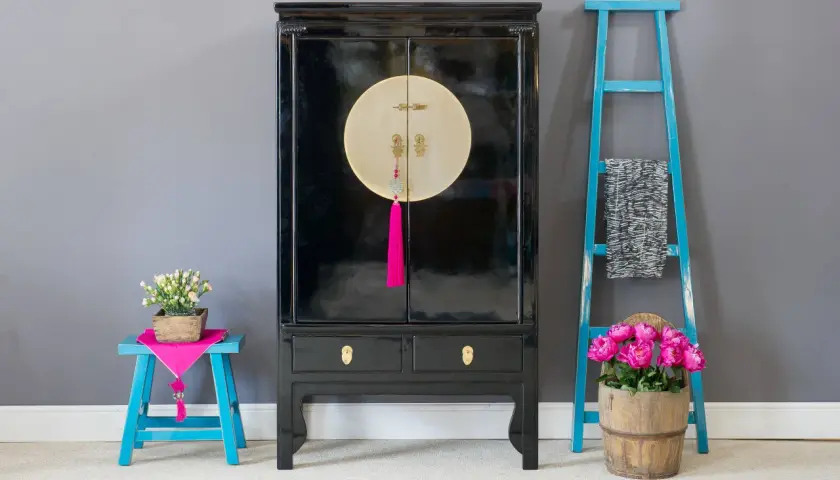 Mobile con laccatura nera lucida, dettagli dorati e nappa fucsia. Accanto, sgabello blu con fiori e scaletta decorativa.