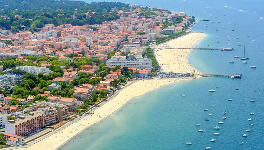 Vue aérienne d'une ville côtière avec une plage bondée et des bateaux dans l'eau.