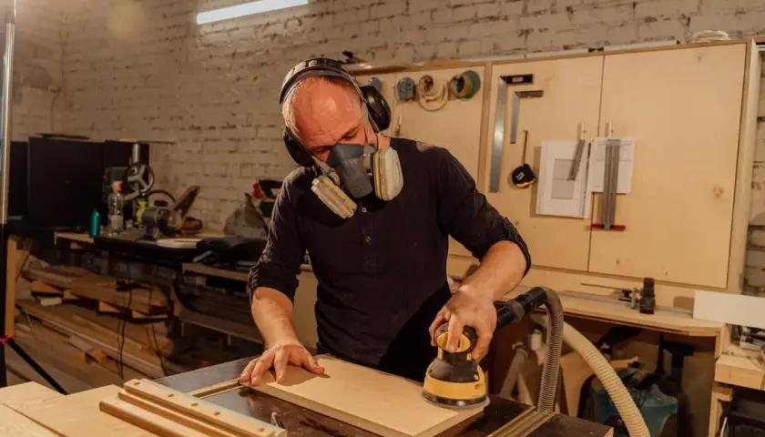 Artisan concentré, masqué et casqué, réalisant un ponçage bois méticuleux avec une ponceuse électrique dans son atelier.