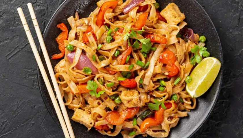 Pad thai z tofu, warzywami i limonką na czarnym talerzu, obok pałeczki.
