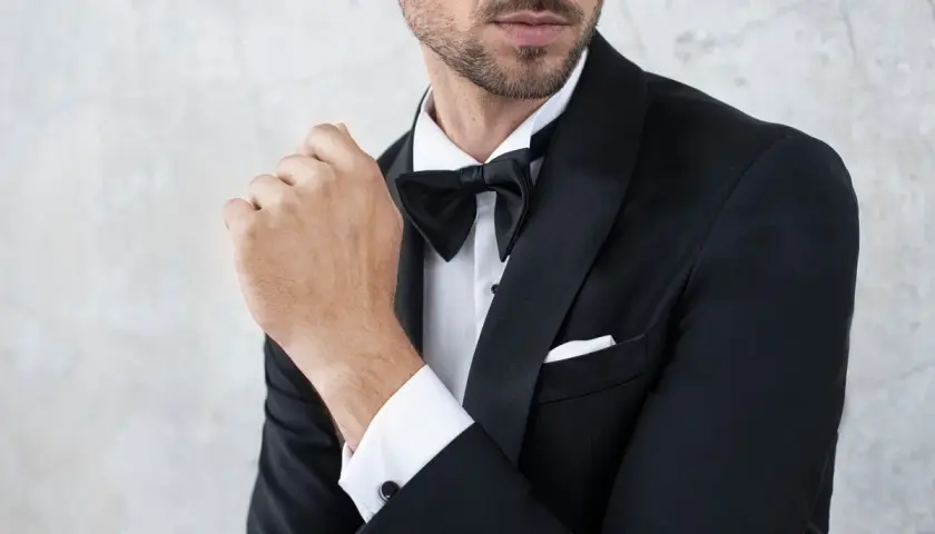 Elegancki mężczyzna w czarnym smokingu i muszce, z dłonią przy ustach.
