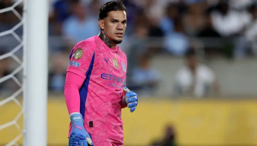 Bramkarz Manchester City w różowym stroju, z niebieskimi rękawicami, gotowy do obrony bramki.