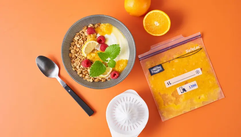 Orangensaft in einem Gefrierbeutel zeigt, dass man orangen einfrieren kann. Daneben ein Müsli mit Früchten.